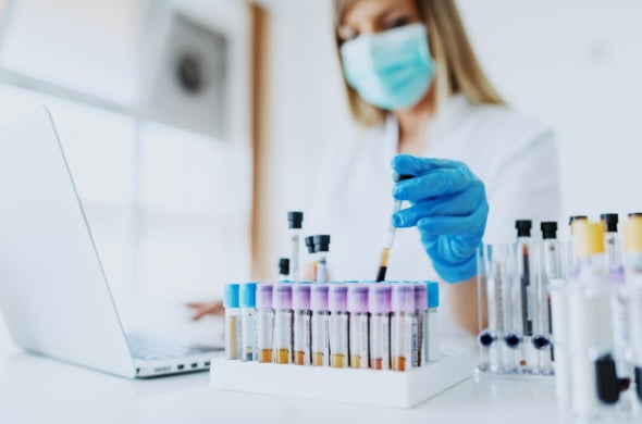 Primer plano del asistente de laboratorio en uniforme, con máscara y guantes de goma sosteniendo el tubo de ensayo con muestra de sangre mientras está sentado en la silla y escribiendo en el ordenador portátil. Enfoque selectivo en tubos de ensayo.